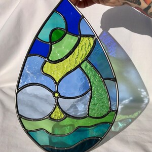 May include: A teardrop-shaped stained glass window with a blue, green, and yellow abstract design. The glass is textured and has a variety of colors.