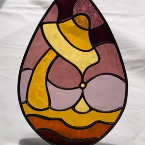 May include: A stained glass teardrop-shaped suncatcher with abstract shapes in shades of purple, pink, and yellow.