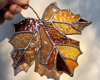 May include: A stained glass maple leaf with a copper wire hanger. The leaf is made of various shades of amber, brown, and red glass. The glass has a textured, antique look.