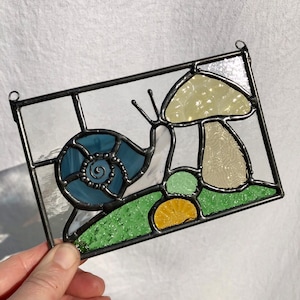 May include: A stained glass window hanging features a snail with a blue shell and a yellow mushroom. The snail is crawling on a green and yellow background.