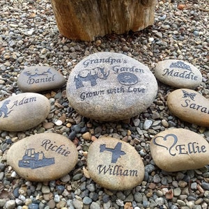 May include: A collection of personalized stones with various designs and names, including "Grandpa's Garden Grown with Love", "Maddie", "Sara", "Adam", "Daniel", "Richie", "William", and "Ellie".