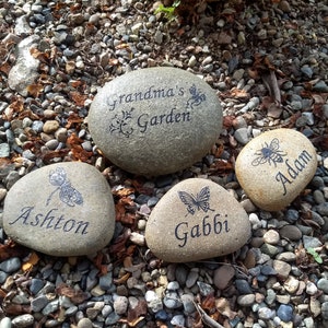 May include: Four engraved stones with different designs and names. The stones are on a bed of small gravel. The stones read "Grandma's Garden", "Ashton", "Gabbi", and "Adam".