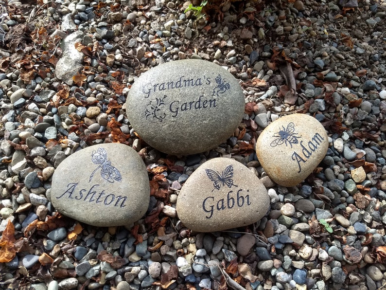 May include: Four engraved stones with different designs and names. The stones are on a bed of small gravel. The stones read "Grandma's Garden", "Ashton", "Gabbi", and "Adam".