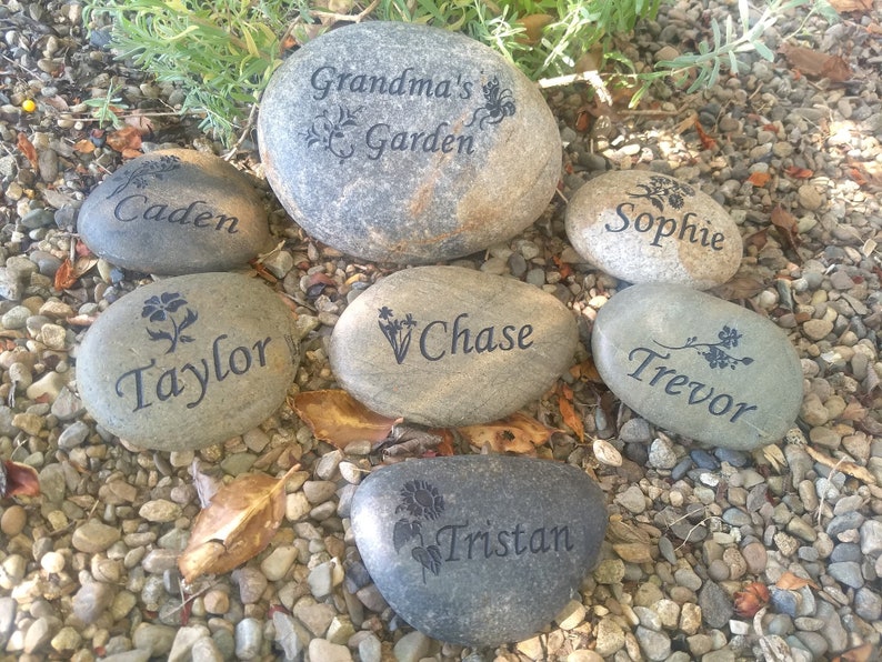 May include: Seven gray river rocks with black engraved text and floral designs. The rocks are labeled "Grandma's Garden", "Caden", "Taylor", "Chase", "Sophie", "Trevor", and "Tristan".