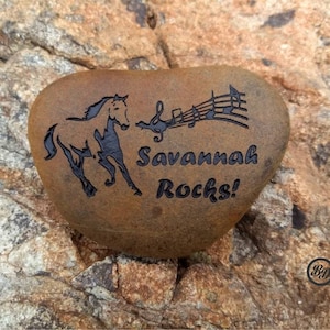 May include: A brown rock with a black silhouette of a horse and musical notes engraved on it. The text "Savannah Rocks!" is also engraved on the rock.