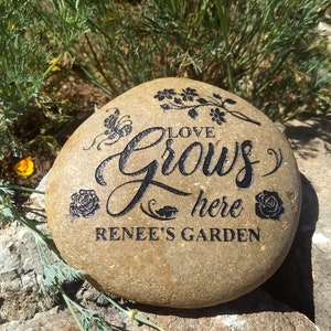 May include: A brown garden stone with the words "Love Grows Here Renee's Garden" engraved in black. The stone is decorated with a butterfly, flowers, and roses.