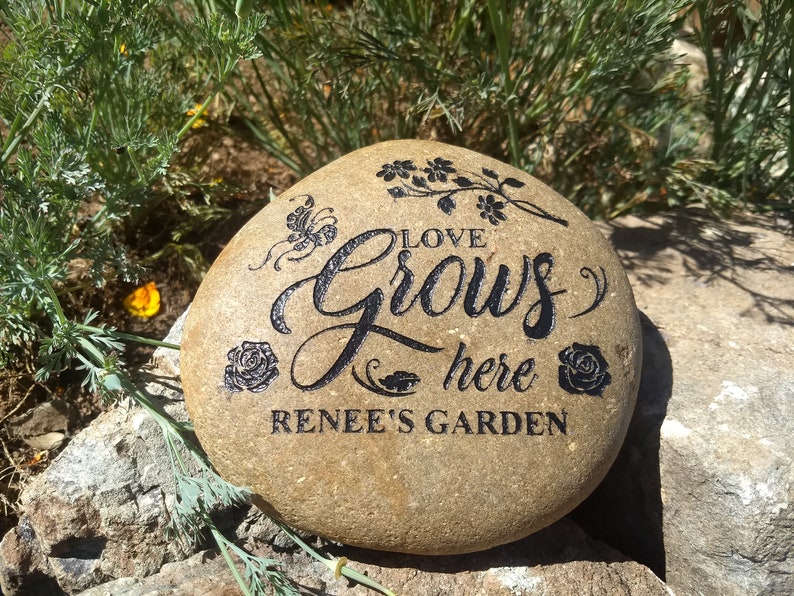 May include: A brown garden stone with the words "Love Grows Here Renee's Garden" engraved in black. The stone is decorated with a butterfly, flowers, and roses.