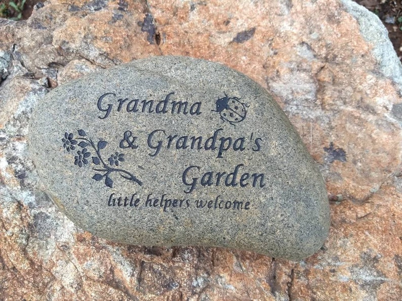 May include: A gray stone with the words "Grandma & Grandpa's Garden little helpers welcome" engraved on it. A small ladybug is also engraved on the stone.