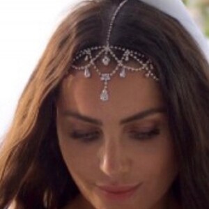 May include: A silver headpiece with clear crystals. The headpiece is worn on the forehead and has a delicate design with dangling crystal accents. The woman has long brown hair and is wearing a white veil.