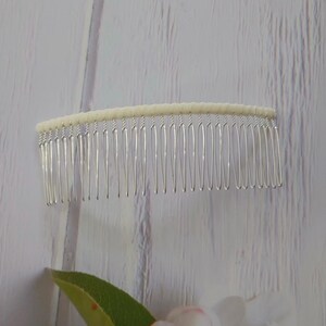 May include: A hair comb featuring a row of thin, silver-toned metal teeth attached to a curved, off-white fabric-wrapped top bar. The design is simple and functional, with the metal teeth extending downward from the textured, light-colored base.