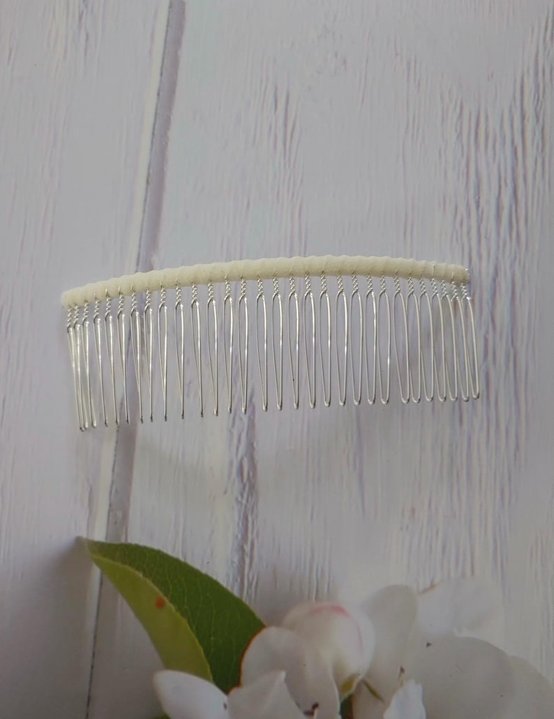 May include: A hair comb featuring a row of thin, silver-toned metal teeth attached to a curved, off-white fabric-wrapped top bar. The design is simple and functional, with the metal teeth extending downward from the textured, light-colored base.