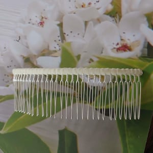 May include: A hair comb featuring a row of thin, silver-toned metal teeth attached to a horizontal bar wrapped in a smooth, pale cream-colored fabric. The design consists of twenty-two individual tines that are evenly spaced and rounded at the tips.
