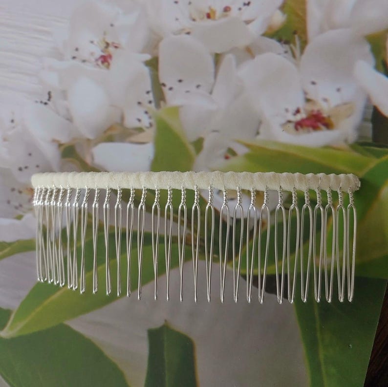 May include: A hair comb featuring a row of thin, silver-toned metal teeth attached to a horizontal bar wrapped in a smooth, pale cream-colored fabric. The design consists of twenty-two individual tines that are evenly spaced and rounded at the tips.