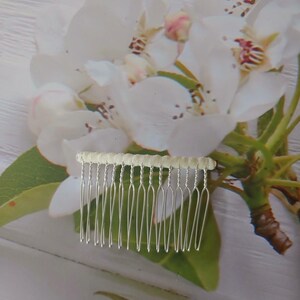 May include: A silver-toned hair comb featuring a row of twelve thin, metallic tines attached to a horizontal bar wrapped in a textured, off-white fabric. The comb is designed with a simple, functional structure for securing hair in place.