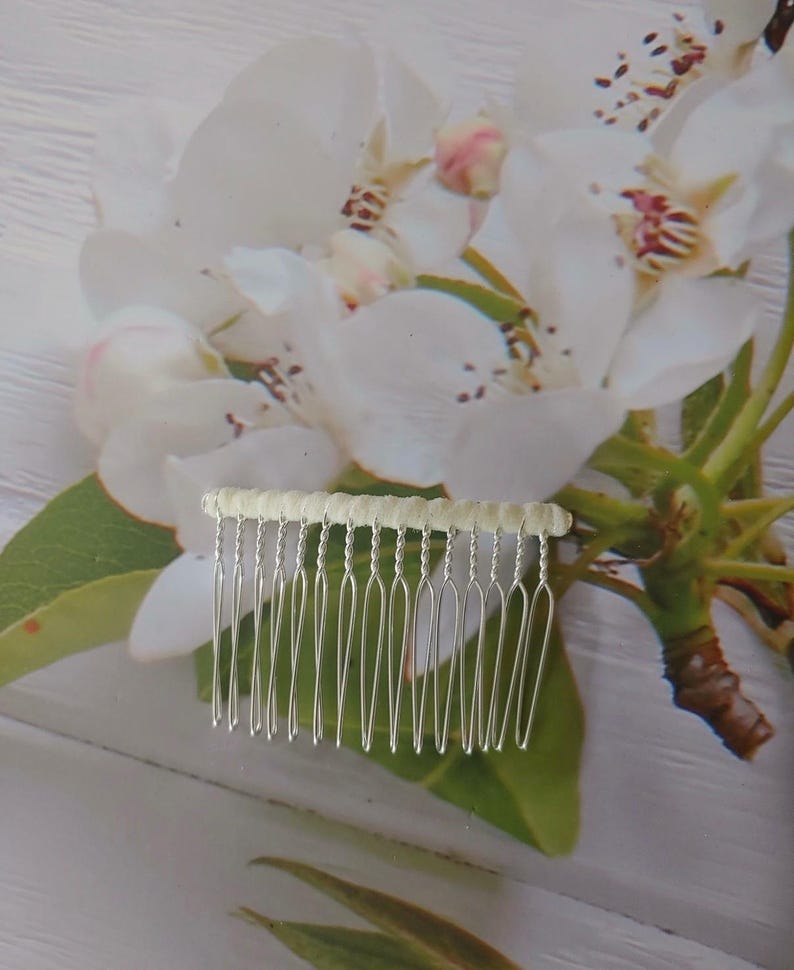 May include: A silver-toned hair comb featuring a row of twelve thin, metallic tines attached to a horizontal bar wrapped in a textured, off-white fabric. The comb is designed with a simple, functional structure for securing hair in place.