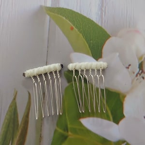 May include: Two hair combs featuring silver-toned metal teeth and a horizontal bar wrapped in off-white fabric. Each comb has a series of thin, vertical tines attached to the fabric-covered top section, designed for securing hair in place.