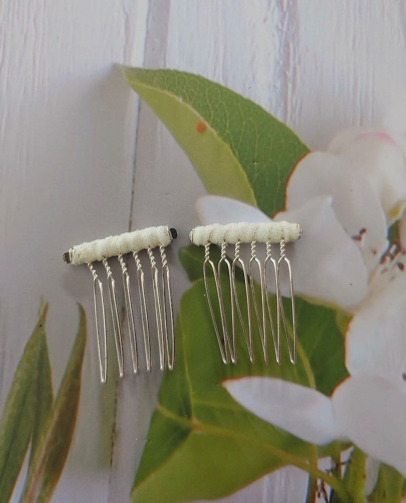May include: Two hair combs featuring silver-toned metal teeth and a horizontal bar wrapped in off-white fabric. Each comb has a series of thin, vertical tines attached to the fabric-covered top section, designed for securing hair in place.