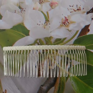 May include: A hair comb featuring a row of thin, silver-toned metal teeth attached to a horizontal bar wrapped in a textured, off-white fabric. The comb has a wide, rectangular shape with evenly spaced, slender prongs designed for securing hair.