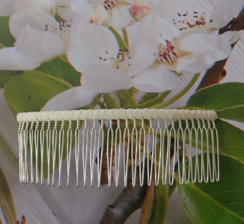 May include: A hair comb featuring a row of thin, silver-toned metal teeth attached to a horizontal bar wrapped in a textured, off-white fabric. The comb has a wide, rectangular shape with evenly spaced, slender prongs designed for securing hair.