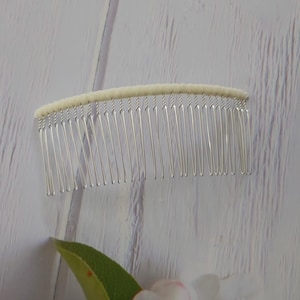 May include: A hair comb featuring a curved, off-white fabric-wrapped top edge and a series of thin, silver-toned metal teeth. The design is simple and functional, with the fabric covering the spine of the comb to provide a clean, finished look.