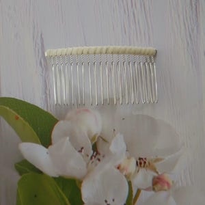 May include: A hair comb featuring a row of thin, silver-toned metal teeth attached to a horizontal bar wrapped in a cream-colored fabric. The comb is positioned above a cluster of white apple blossoms with green leaves and pink-tipped petals.