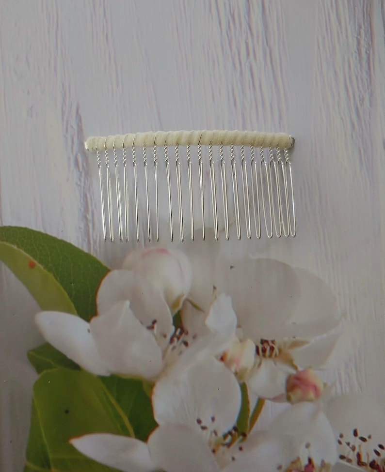 May include: A hair comb featuring a row of thin, silver-toned metal teeth attached to a horizontal bar wrapped in a cream-colored fabric. The comb is positioned above a cluster of white apple blossoms with green leaves and pink-tipped petals.