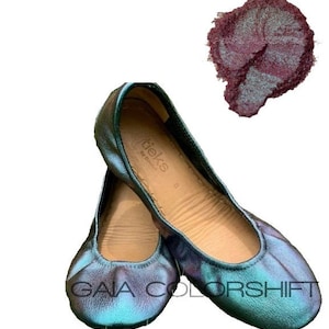 May include: A pair of teal green ballet flats with a color-shifting finish. The flats are made of leather and have a rounded toe. The brand name "tieks" is printed on the insole. The flats are shown next to a sample of teal green shimmery pigment.
