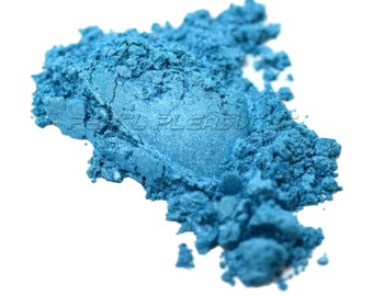 May include: A pile of loose, vibrant blue mica powder with a shimmering, metallic finish. The powder has a fine, granular texture and is arranged in an irregular mound. The text PEARL PLEASURES is visible across the center of the powder.