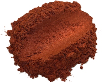 May include: A pile of reddish-brown powder with a metallic sheen. The powder has a fine, granular texture and appears to be a pigment or cosmetic product. The color is a warm, earthy tone, possibly a shade of copper or bronze.