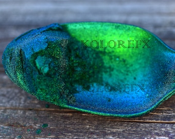 May include: A scoop filled with fine, iridescent pigment powder that shifts in color from deep blue to vibrant green. The powder has a sparkling, metallic texture. The text KOLOREFX is visible in black lettering across the surface of the powder.