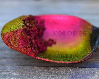 May include: A black spoon filled with vibrant glitter. The glitter transitions from green to pink, with the text "KOLOREFX" visible. The spoon is set against a weathered wooden surface.
