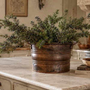 May include: A round, dark brown metal bucket with two side handles, filled with green juniper branches. The bucket sits on a light-colored stone countertop next to a wooden bowl placed on top of two stacked books, one titled Maison Rustique.