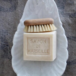 May include: A square block of beige Savon de Marseille soap with the text embossed on the front, topped with a small wooden scrub brush featuring light-colored bristles, all resting on a white ceramic dish shaped like a scalloped shell.