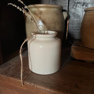 May include: A small, off-white ceramic jar with a cylindrical body and a short neck. The base of the jar features the embossed text "AB Lagny". A piece of twine is tied around the neck, holding two dried lavender stems inside the opening.