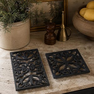 May include: Two black cast iron trivets with intricate scrollwork patterns. One trivet is rectangular in shape, while the other is square. Both feature openwork designs with swirling metal details, providing a stable base for hot cookware.