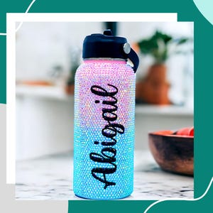 May include: A blue and pink ombre water bottle with a black lid and a black strap. The bottle is covered in rhinestones and has the name "Abigail" written on it in black.