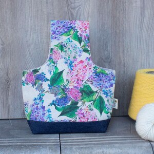 May include: A fabric yarn bag with a floral pattern of purple, pink, and blue flowers and green leaves. The bag has a dark blue base and a handle. A yellow yarn cone and white yarn are next to the bag.