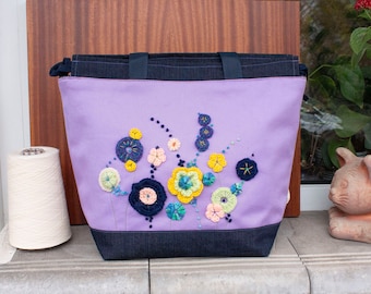 May include: A purple tote bag with a navy blue trim and embroidered flowers in shades of yellow, blue, and green.