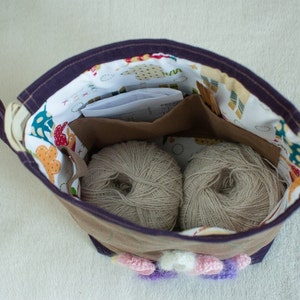 May include: A purple fabric bag with a white and colorful patterned lining. The bag is open and contains two balls of beige yarn.