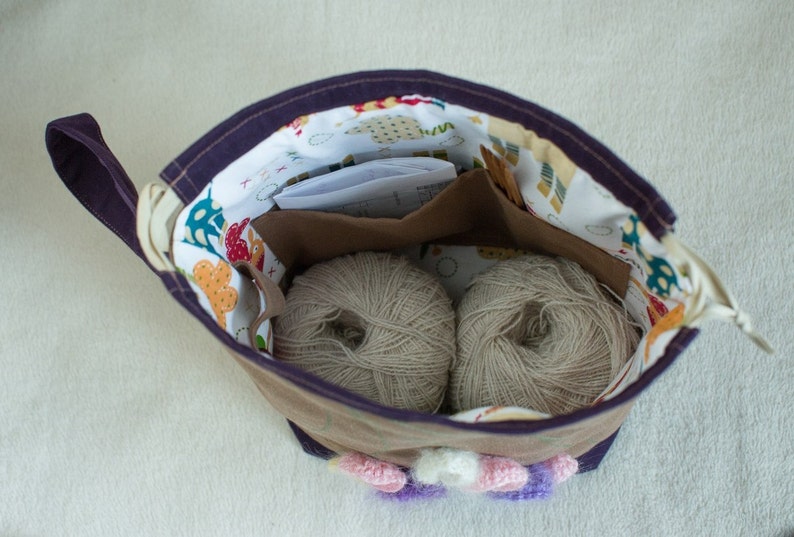 May include: A purple fabric bag with a white and colorful patterned lining. The bag is open and contains two balls of beige yarn.