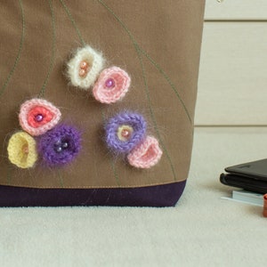 May include: A brown fabric tote bag with a purple trim and crocheted flowers in shades of pink, white, and purple.
