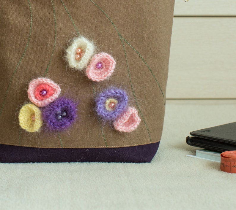 May include: A brown fabric tote bag with a purple trim and crocheted flowers in shades of pink, white, and purple.