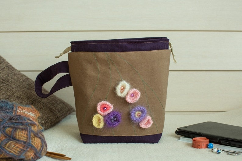 May include: A brown canvas bag with a purple lining and a drawstring closure. The bag has a purple handle and is decorated with crocheted flowers in shades of pink, purple, and yellow.