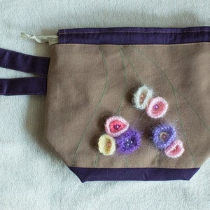May include: A brown canvas bag with a purple handle and a purple trim. The bag has a design of green lines and is decorated with crocheted flowers in shades of pink, white, and purple.