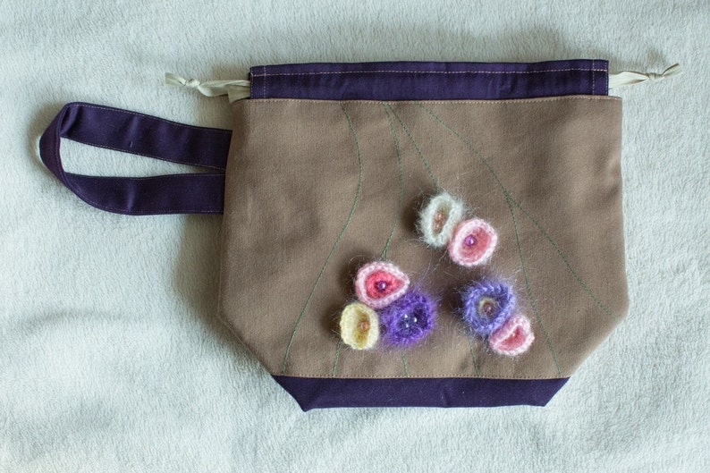 May include: A brown canvas bag with a purple handle and a purple trim. The bag has a design of green lines and is decorated with crocheted flowers in shades of pink, white, and purple.