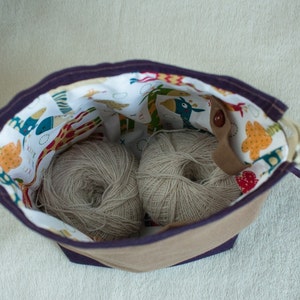 May include: A purple and brown fabric bag with a white lining featuring a colorful animal print. The bag is open and contains two balls of beige yarn.