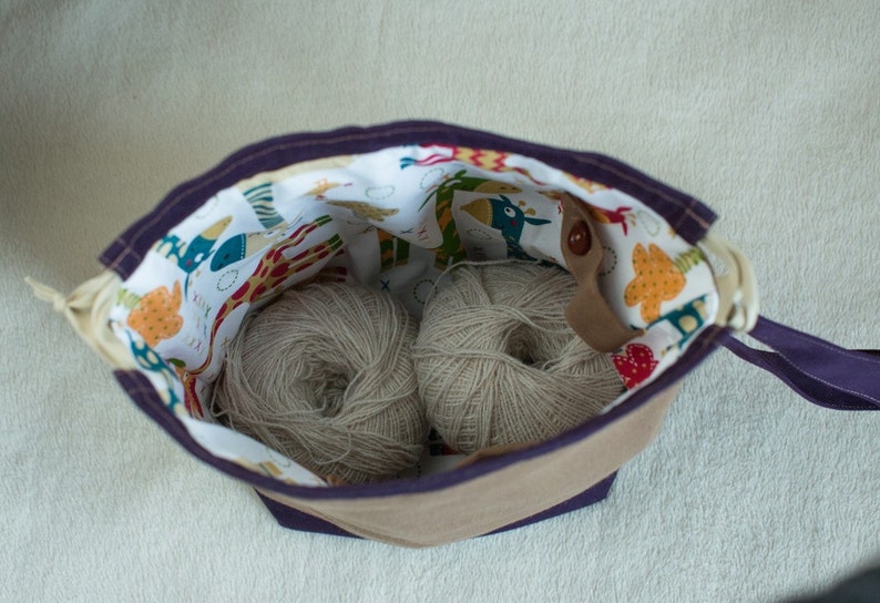 May include: A purple and brown fabric bag with a white lining featuring a colorful animal print. The bag is open and contains two balls of beige yarn.