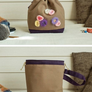 May include: A brown drawstring bag with a purple trim and crocheted flower embellishments. The bag has a purple strap.