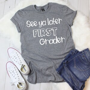 May include: Gray t-shirt with white text that reads "See ya later FIRST Grader". The shirt is laying on a white fuzzy surface with a pair of white sneakers and blue denim shorts.