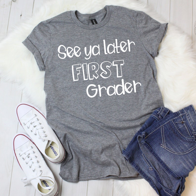 May include: Gray t-shirt with white text that reads "See ya later FIRST Grader". The shirt is laying on a white fuzzy surface with a pair of white sneakers and blue denim shorts.